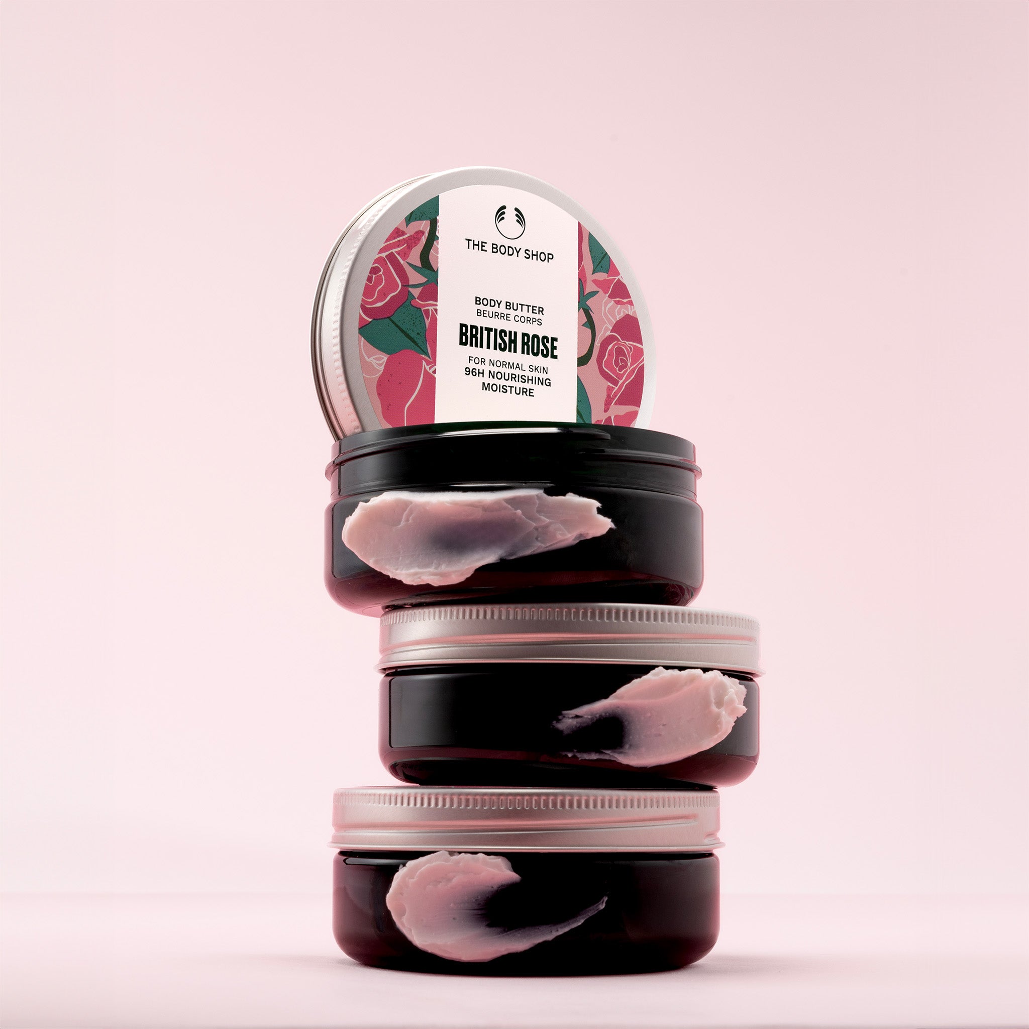 A stack of three The Body Shop British Rose Body Butter tubs with a pink background.