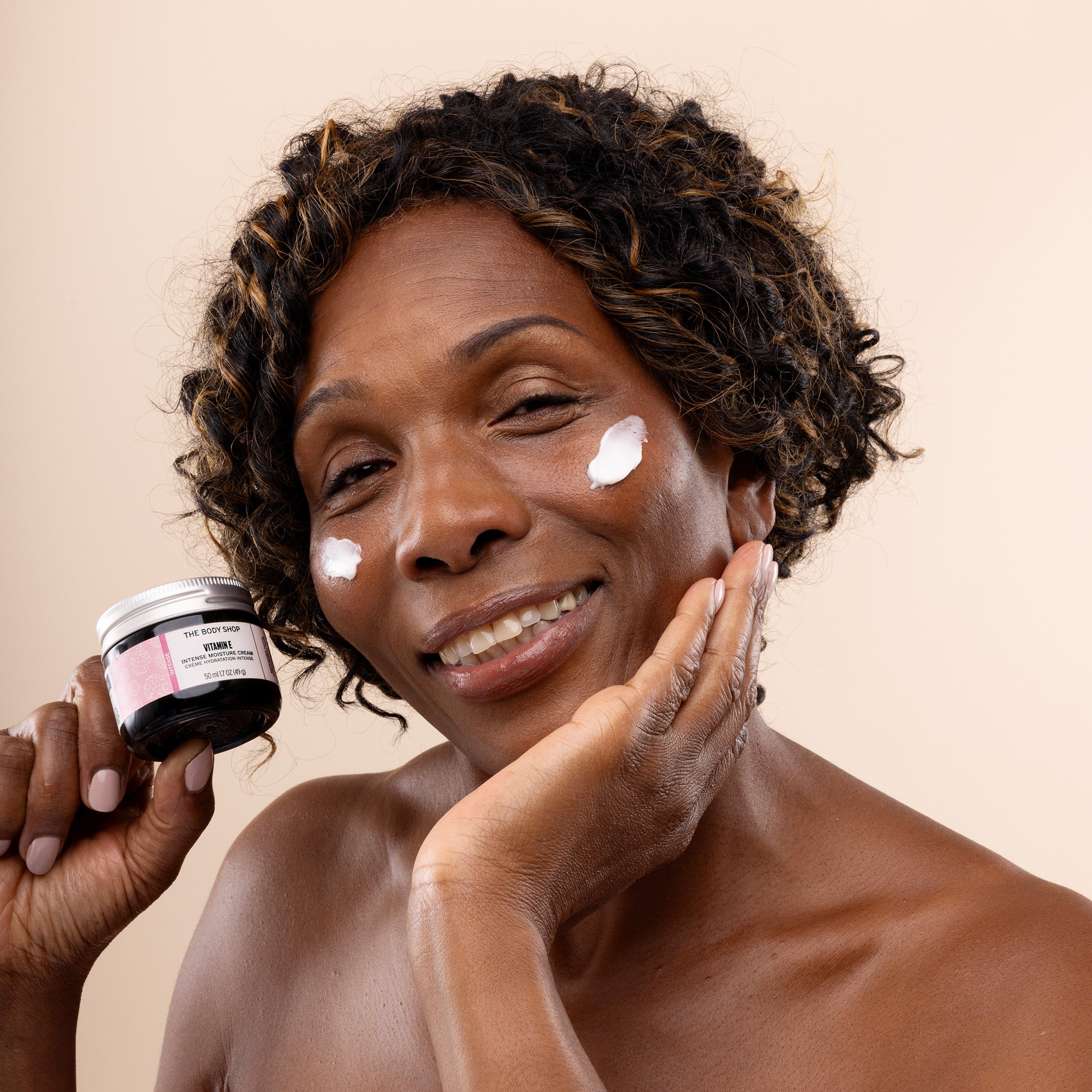 A woman holding a jar of Vitamin E intense moisture cream with two smears of the product on her face.
