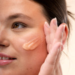 Woman applying The Body Shop Vitamin C Glow Boosting Moisturiser to her cheek with a neutral background