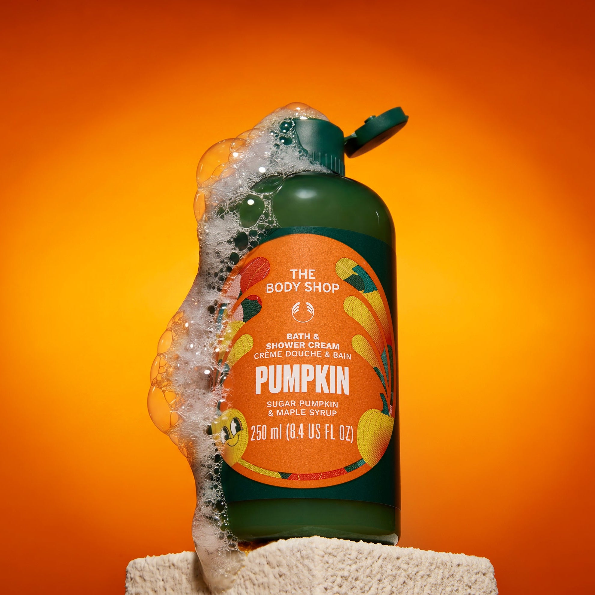 A bottle of The Body Shop Pumpkin bath and shower cream stands on a textured white surface against a vibrant orange gradient background. A stream of white foam is spraying from the open bottle, which features a dark green body and a prominent orange label with illustrations of pumpkins.