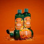 Three bottles of The Body Shop Pumpkin Body & Sweatshirt Care are arranged on a bright orange background with clear bubbles around them. Two bottles stand upright, and one lies on its side in front.