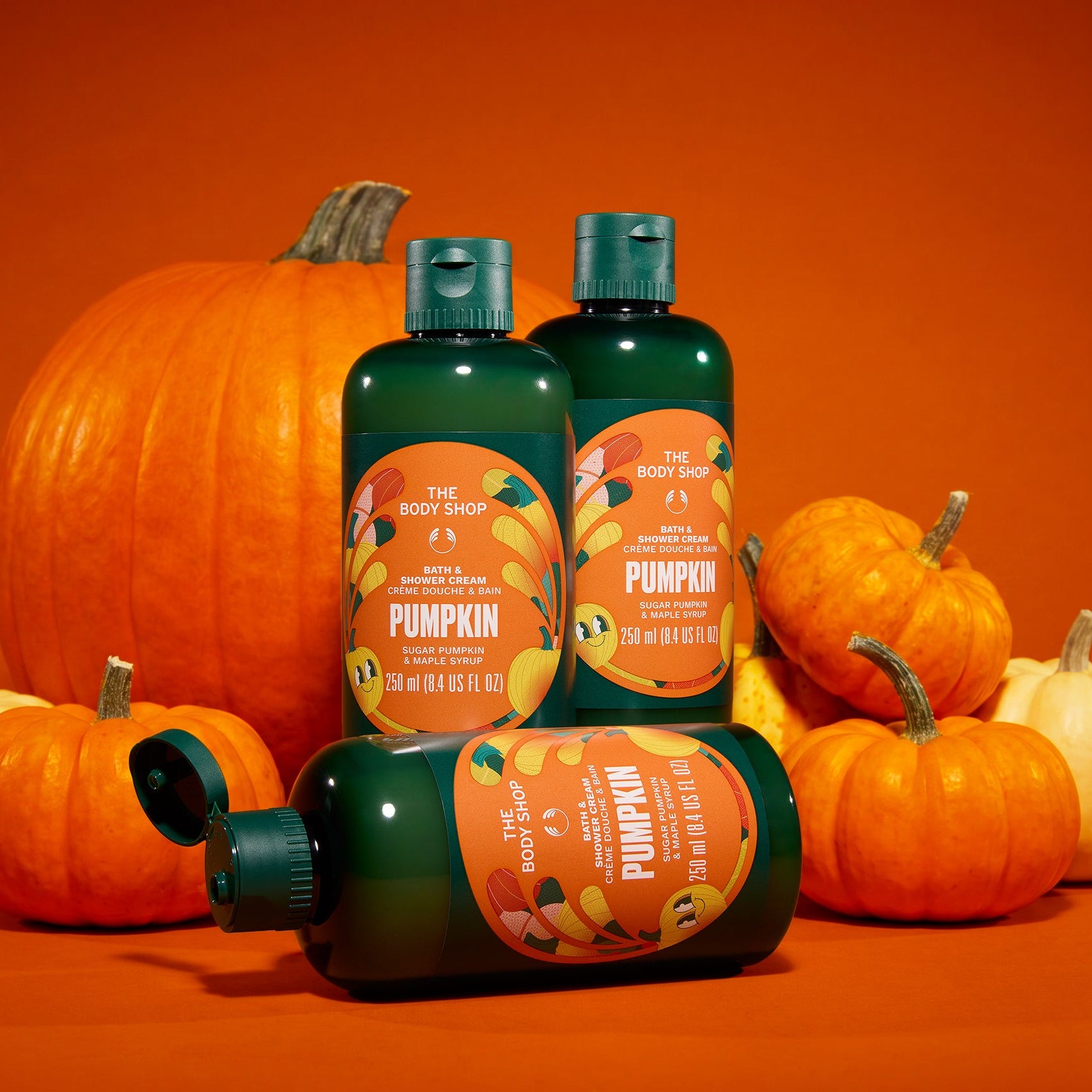 Three bottles of The Body Shop Pumpkin Shea & Shea Cream body wash arranged on a bright orange surface with pumpkins around them. Two bottles stand upright, and one lies on its side in front.