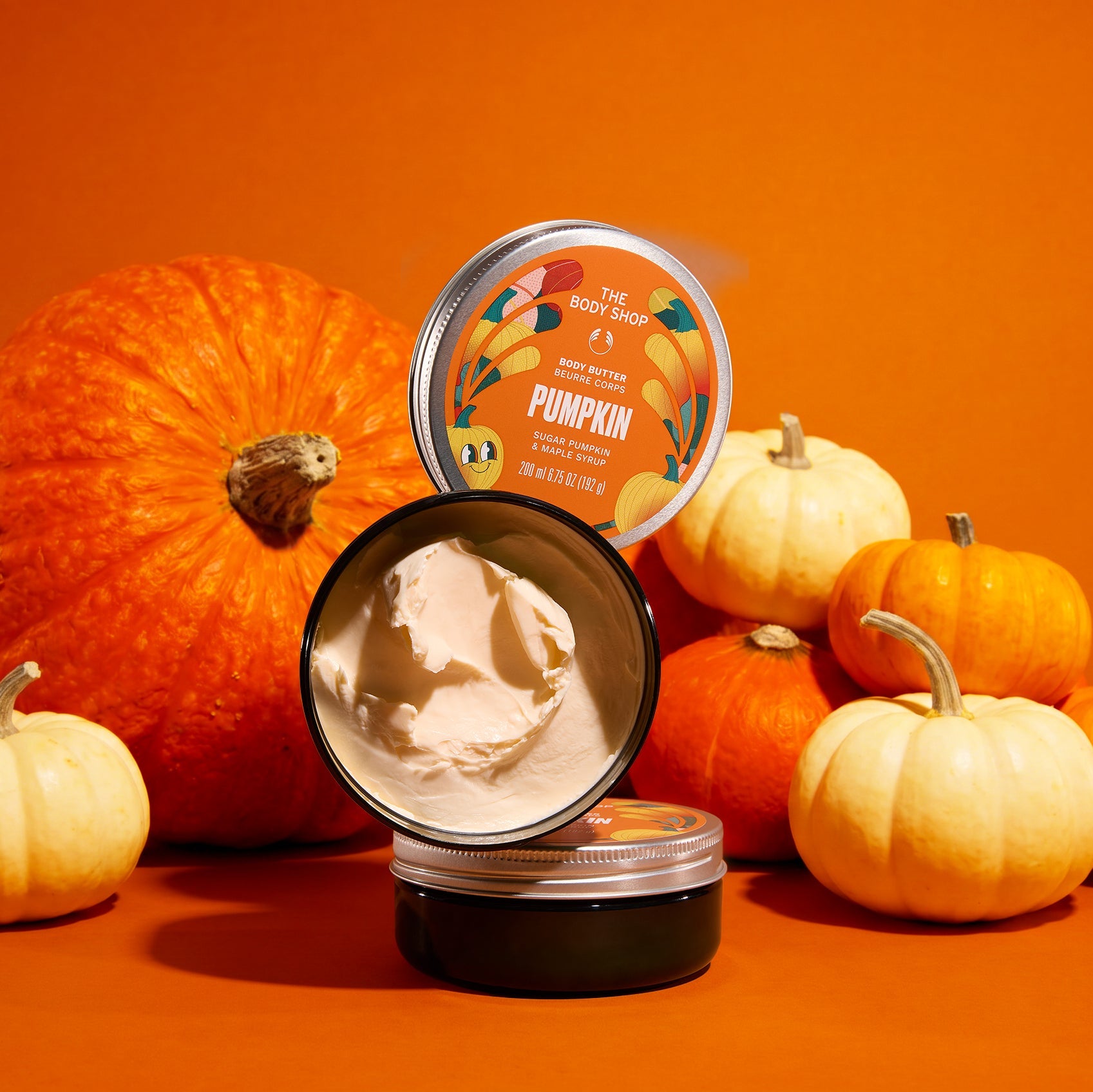 A body butter product tin shown open with cream-colored body butter inside, surrounded by pumpkins on an orange studio background. The lid is propped behind the tin, and the label reads 'The Body Shop' and 'Pumpkin' with autumn-themed illustrations.