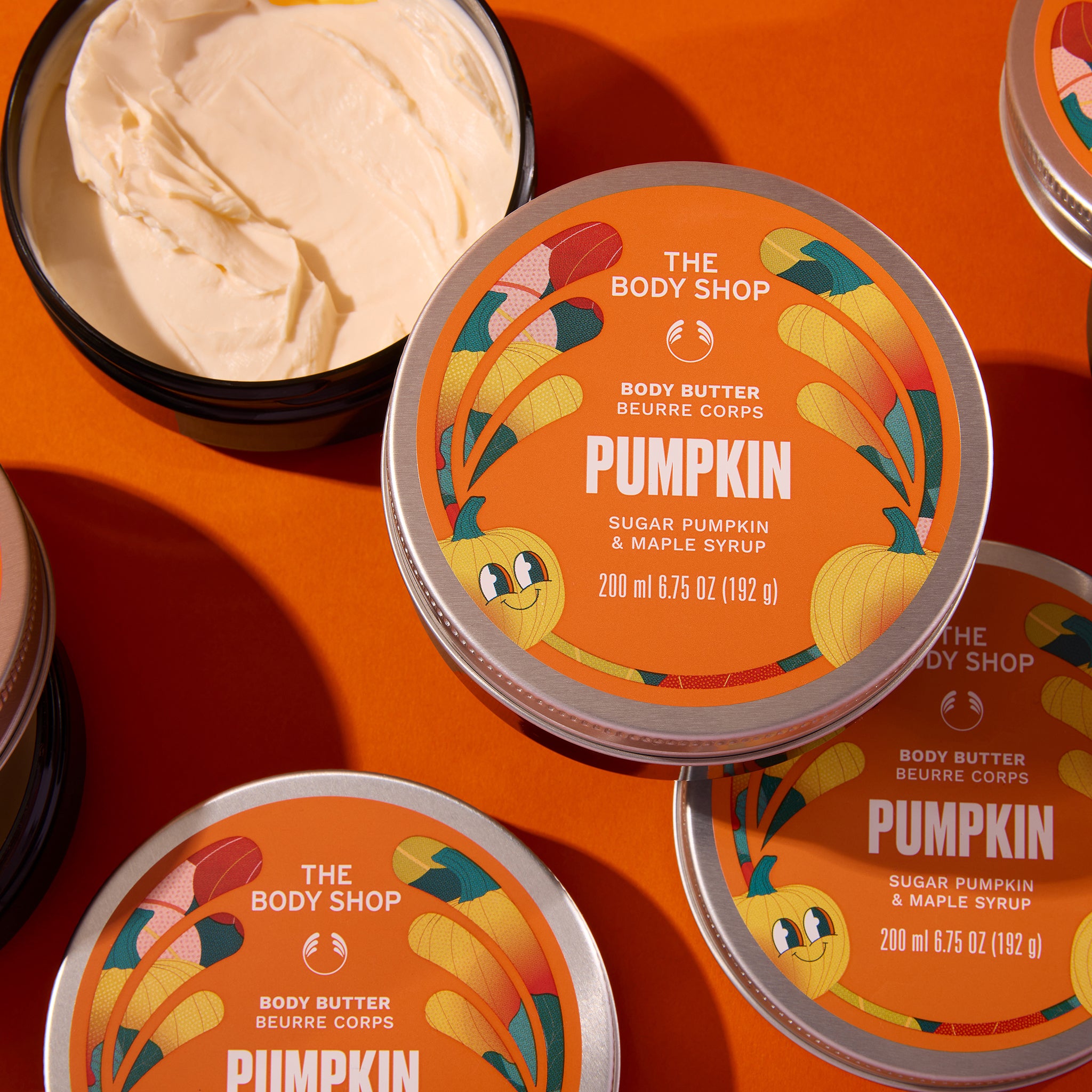 Three round tins of The Body Shop Pumpkin Body Butter arranged on a warm orange background. One tin is open, showing the creamy off-white body butter inside.