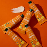 Five tubes of The Body Shop Hand Balm Magic Wand Pumpkin scent arranged diagonally on a bright orange background, with a white cream smear above them.