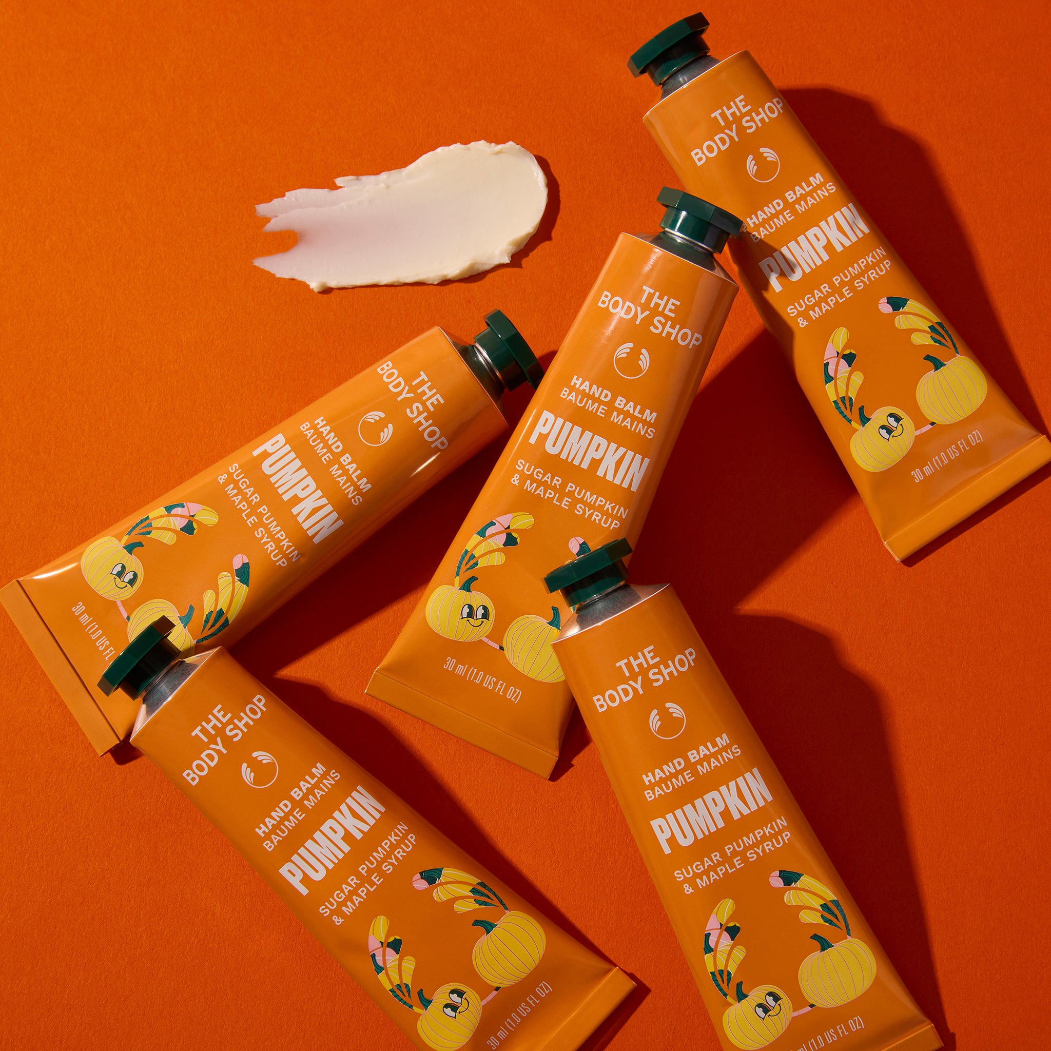 Five tubes of The Body Shop Hand Balm Magic Wand Pumpkin scent arranged diagonally on a bright orange background, with a white cream smear above them.