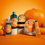 A set of The Body Shop 'Pumpkin' scented products arranged on a wooden pedestal against a bright orange background with decorative pumpkins. The products include a clear spray bottle, a dark green lotion bottle, a dark jar, a round tub, and a yellow hand cream tube.