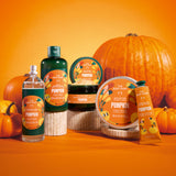 A set of The Body Shop 'Pumpkin' scented products arranged on a wooden pedestal against a bright orange background with decorative pumpkins. The products include a clear spray bottle, a dark green lotion bottle, a dark jar, a round tub, and a yellow hand cream tube.