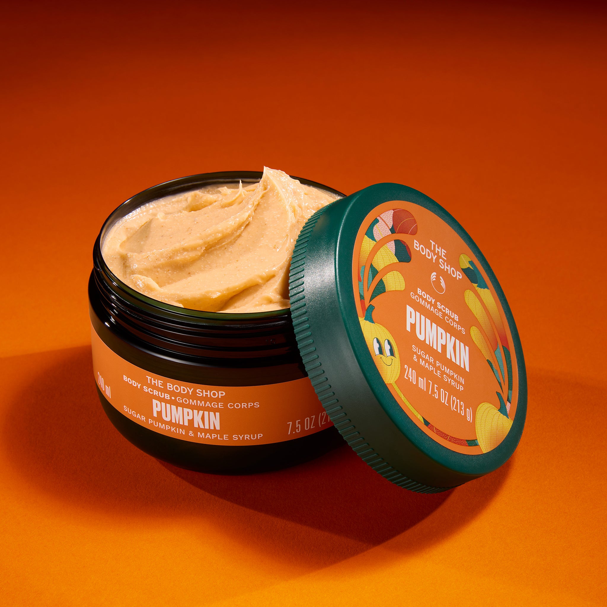 An open jar of The Body Shop Pumpkin Body Butter with its lid leaning against it, set against a solid orange background. The body butter is a light pumpkin-colored cream, and the jar features a dark green lid with a matching orange label.