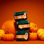 Three dark green cosmetic jars labelled Pumpkin are stacked in a pyramid in front of several orange pumpkins on a solid orange background.