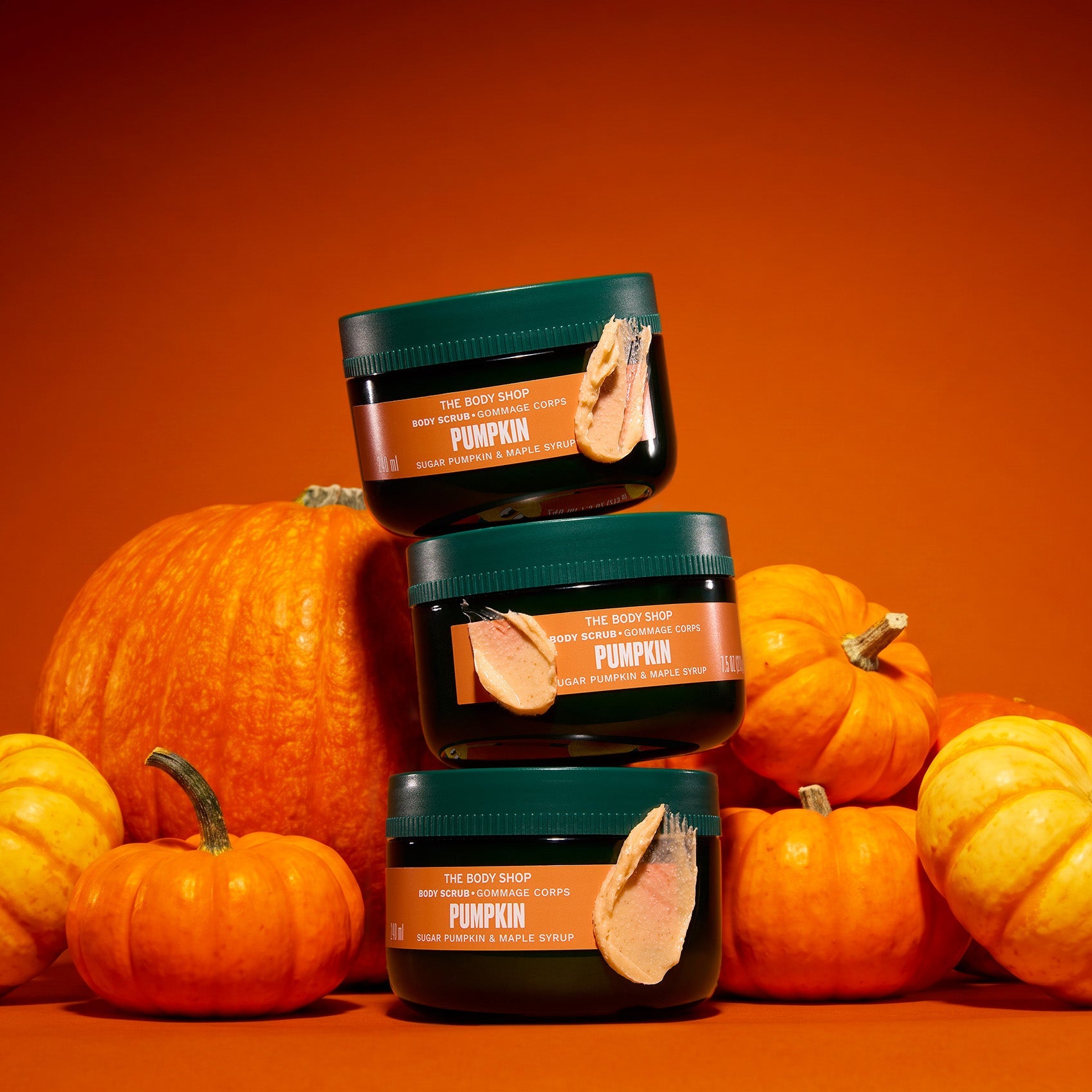 Three dark green cosmetic jars labelled Pumpkin are stacked in a pyramid in front of several orange pumpkins on a solid orange background.
