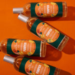 Four bottles of The Body Shop Pumpkin body wash arranged diagonally on a vibrant orange background. Each clear bottle contains a light peach-colored liquid and is topped with a silver cap, featuring a dark green and orange label with illustrations of pumpkins and text.