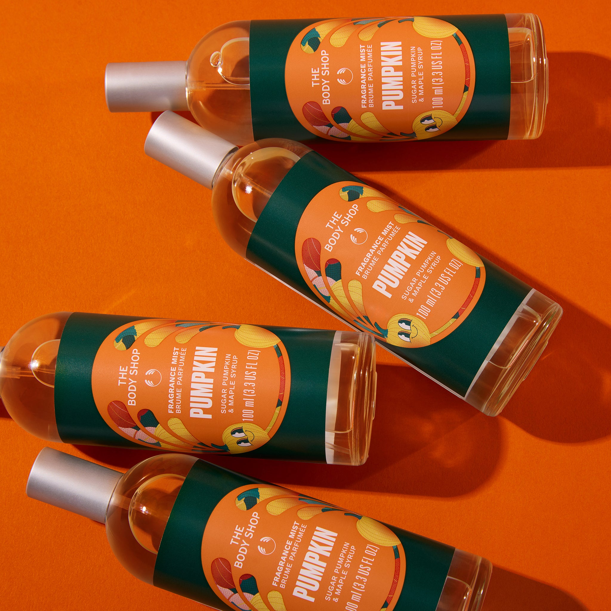 Four bottles of The Body Shop Pumpkin body wash arranged diagonally on a vibrant orange background. Each clear bottle contains a light peach-colored liquid and is topped with a silver cap, featuring a dark green and orange label with illustrations of pumpkins and text.