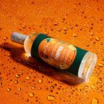A bottle of Scented Flowers 'Pumpkin' fragrance oil lies diagonally on a glossy orange surface covered in water droplets. The bottle has a dark green label with orange and yellow pumpkin illustrations and a silver cap.