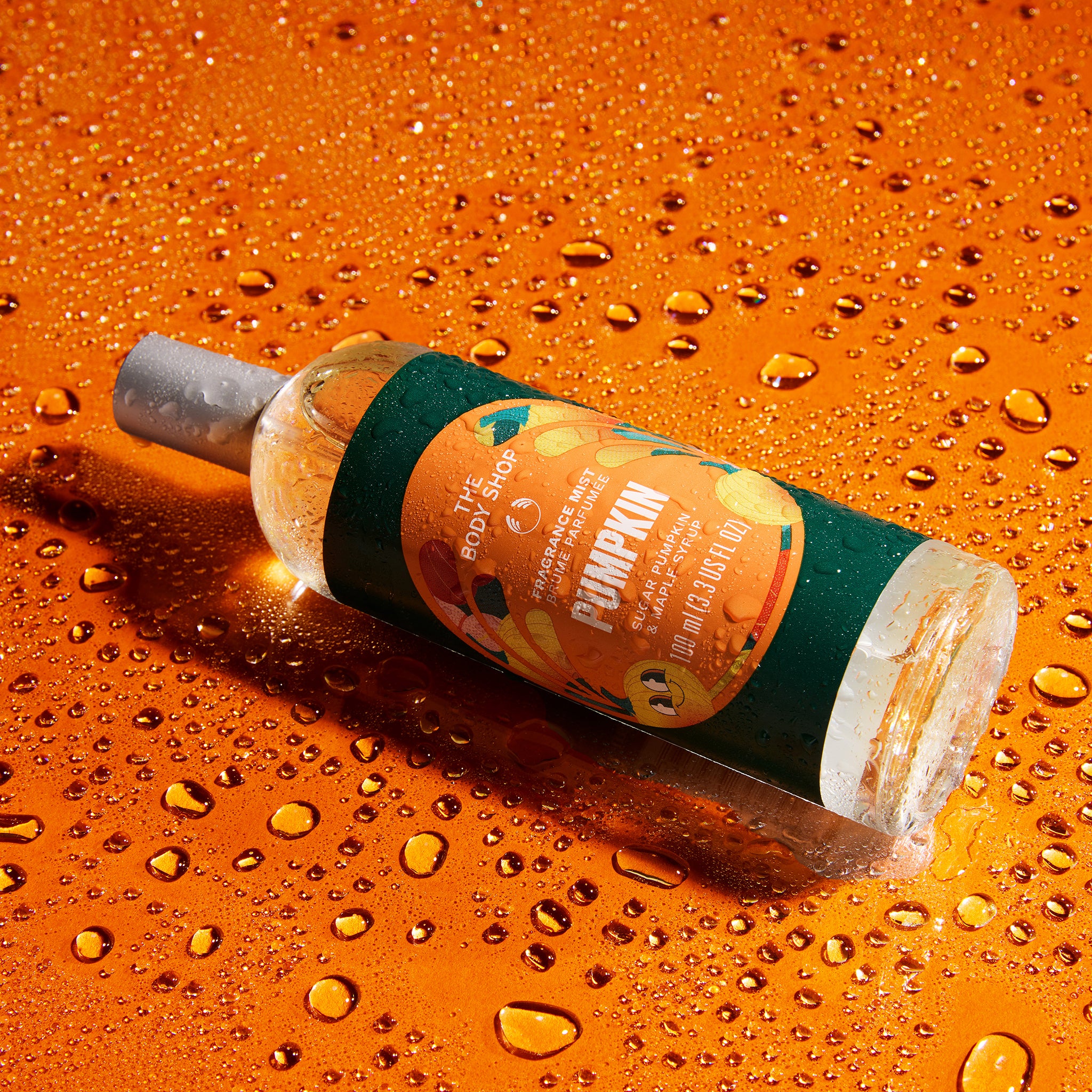 A bottle of Scented Flowers 'Pumpkin' fragrance oil lies diagonally on a glossy orange surface covered in water droplets. The bottle has a dark green label with orange and yellow pumpkin illustrations and a silver cap.