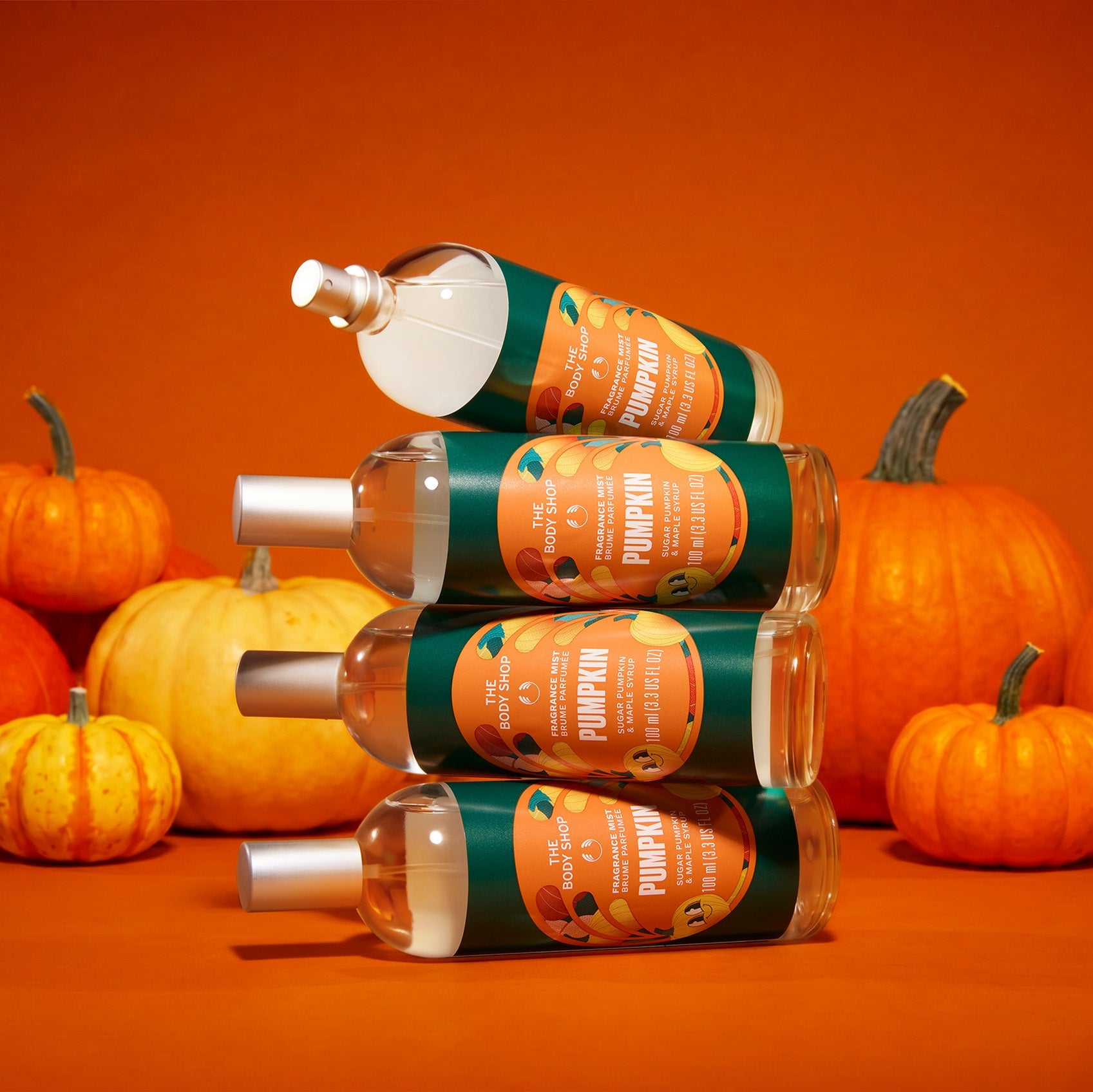 Four bottles of pumpkin spice hair oil arranged in a pyramid stack on an orange background, surrounded by various pumpkins. The bottles have dark green labels with orange illustrations of pumpkins and silver caps.