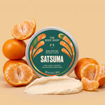 Satsuma body butter jar surrounded by satsumas, with a scoop of cream in front on a light orange background | The Body Shop