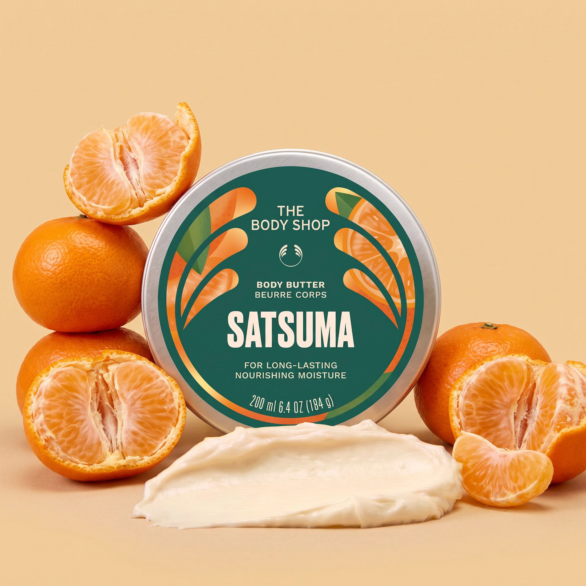 Satsuma body butter jar surrounded by satsumas, with a scoop of cream in front on a light orange background | The Body Shop