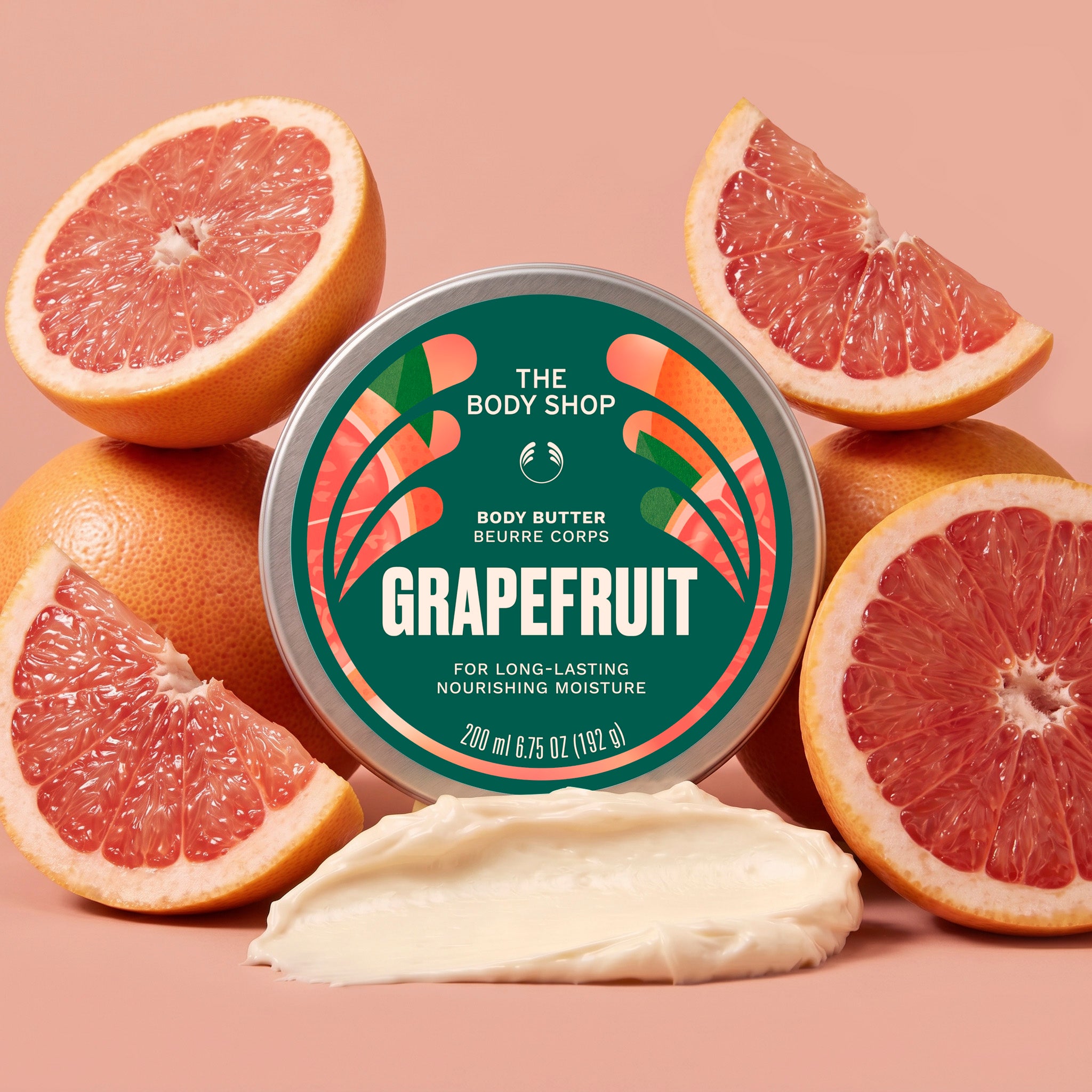 Grapefruit body butter jar surrounded by grapefruits, with a scoop of cream in front on a light brown background | The Body Shop