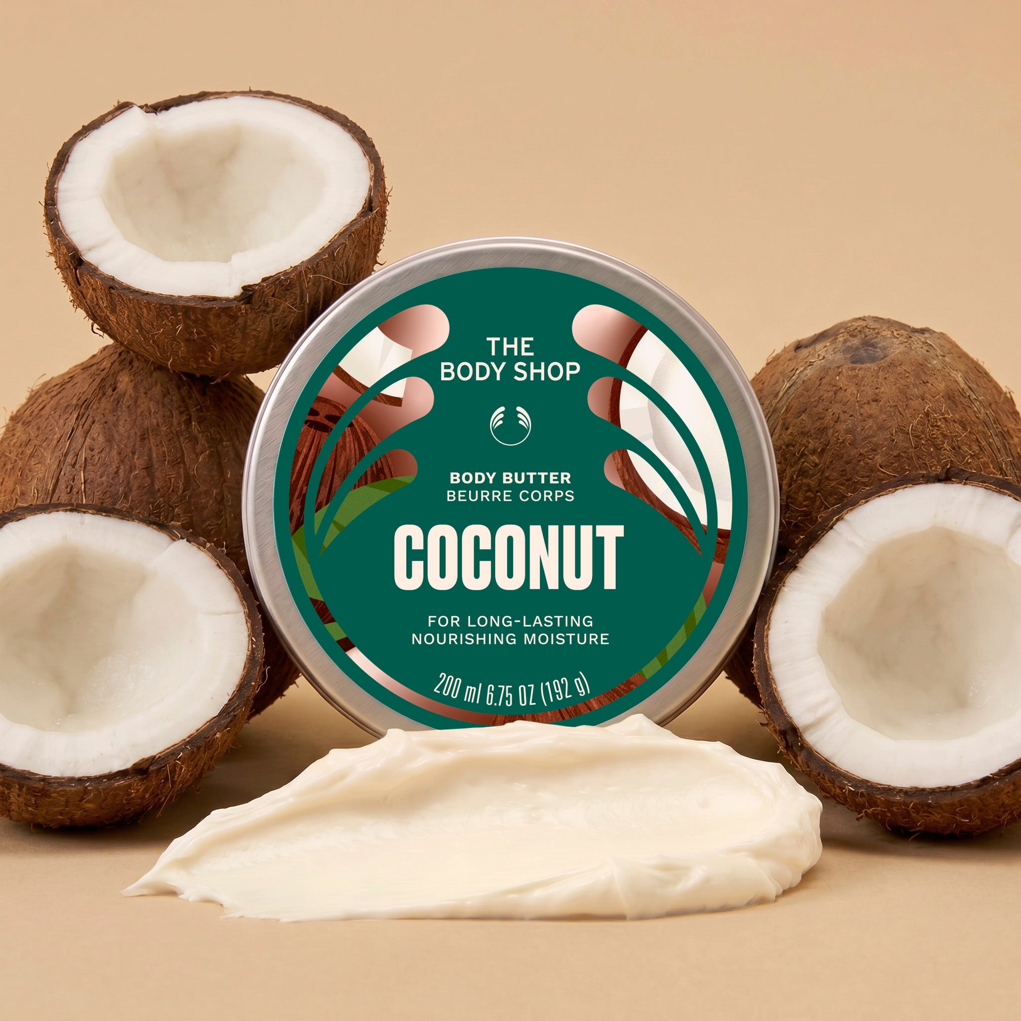 Coconut body butter jar surrounded by coconuts, with a scoop of cream in front on a light brown background | The Body Shop
