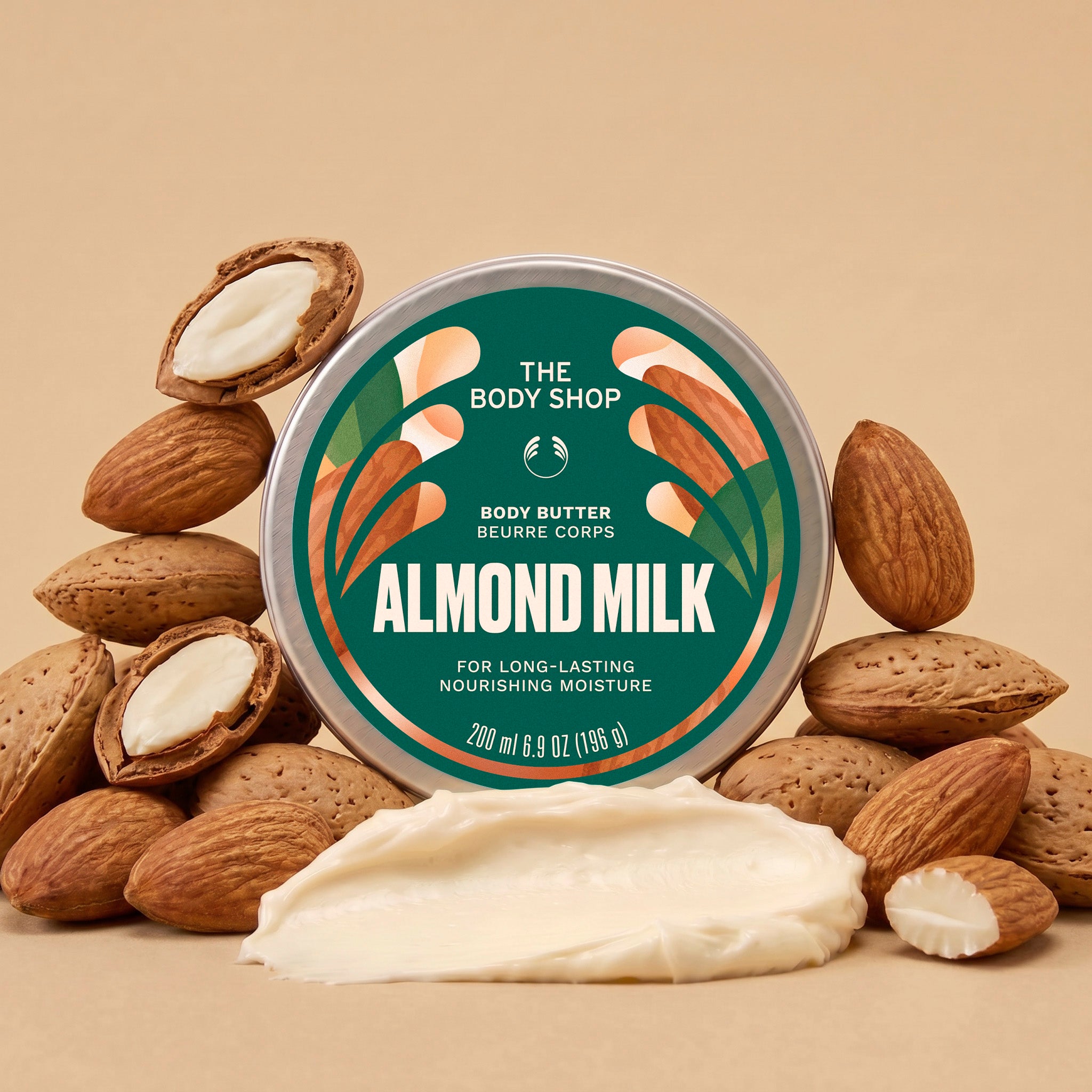 Almond milk body butter jar surrounded by green leaves and white flowers, with a scoop of cream in front on a warm brown background | The Body Shop
