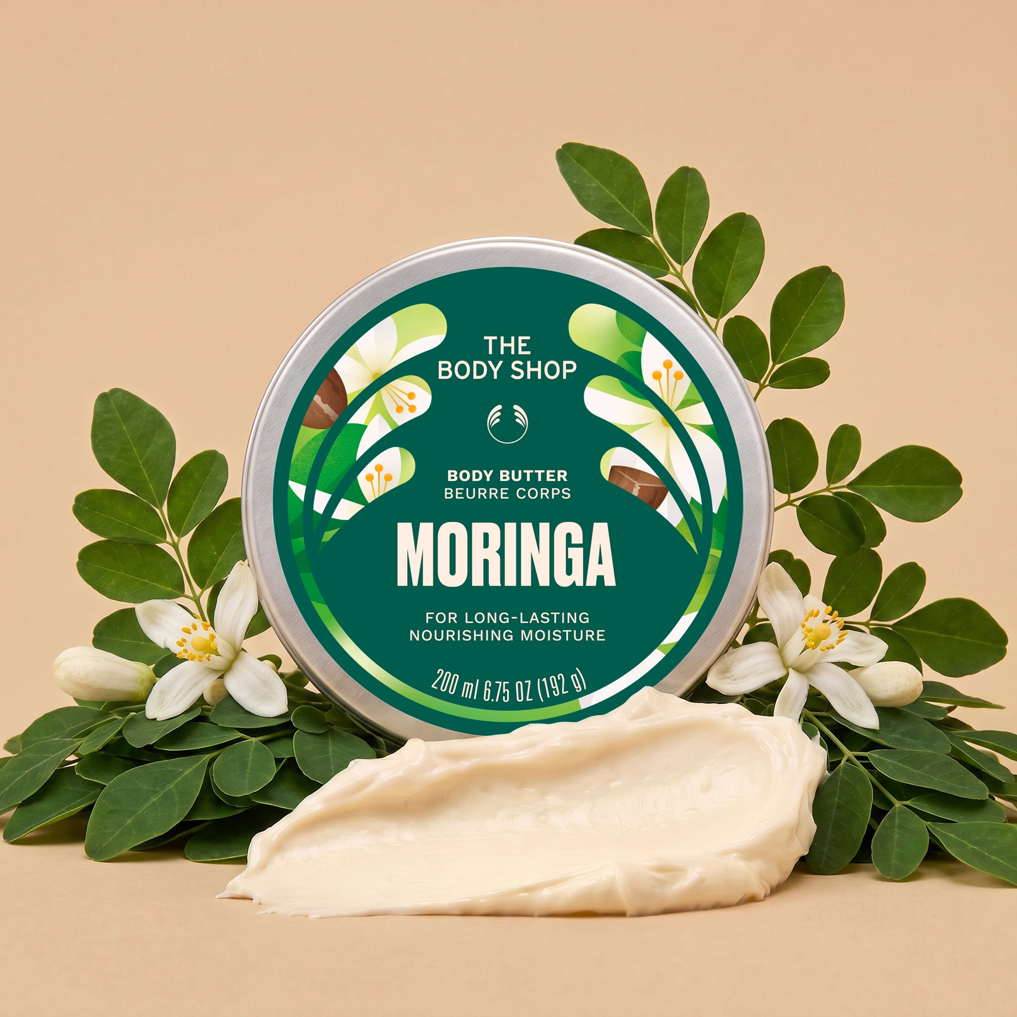 Moringa body butter jar surrounded by green leaves and white flowers, with a scoop of cream in front on a warm beige background | The Body Shop
