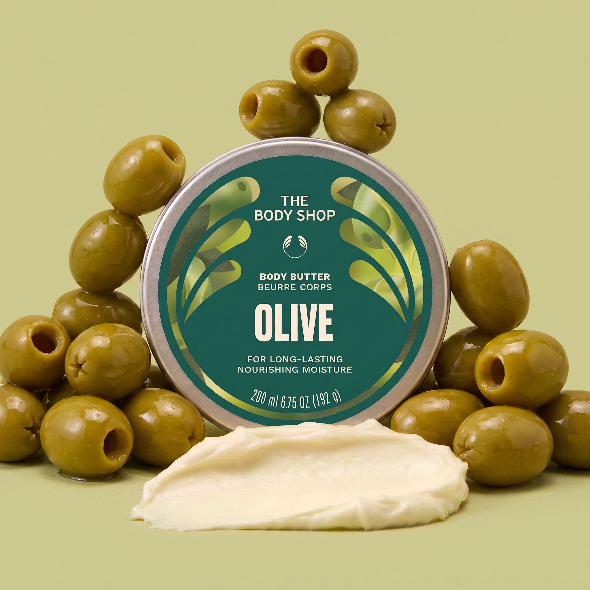 Olive body butter jar surrounded by olives, with a scoop of cream in front on an olive green background | The Body Shop
