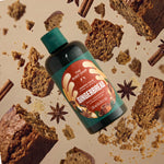 A bottle of The Body Shop Gingerbread room spray is shown floating among gingerbread slices, crumbs, cinnamon sticks, and star anise on a warm beige background.