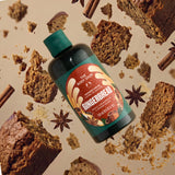A bottle of The Body Shop Gingerbread room spray is shown floating among gingerbread slices, crumbs, cinnamon sticks, and star anise on a warm beige background.