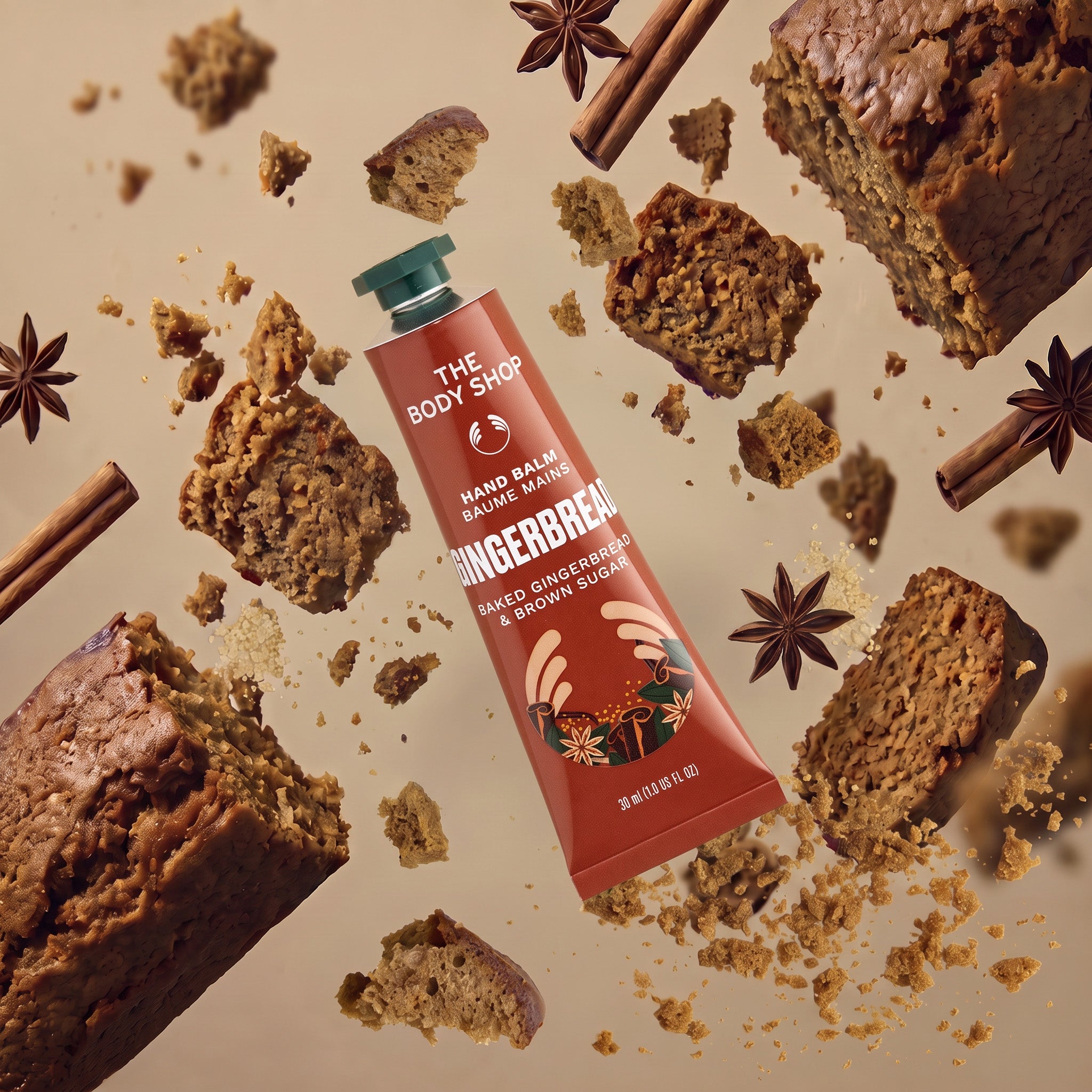 A squeeze tube of 'The Body Shop' Gingerbread Hand Balm is centered in a dynamic flat-lay composition surrounded by gingerbread slices, crumbs, cinnamon sticks, and star anise on a warm beige background.