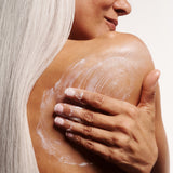 Woman applying Shea Body Butter to her shoulder and back with a beige background