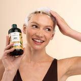 Woman with soapy hair holding a bottle of shampoo against a beige background - The Body Shop Ginger Anti-dandruff Shampoo